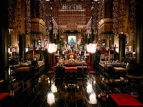 An ornate interior of a traditional temple featuring intricate wooden carvings and golden decorations. The space is filled with various ritual objects, including lanterns, cushions, and altars adorned with offerings. The dim lighting creates a serene and sacred atmosphere.
