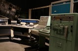A vintage computer room featuring large, retro computers and control panels with numerous buttons and switches. The room includes a UNIVAC 1232 machine and several other large computing devices, arranged in a dimly lit space that emphasizes their mechanical and industrial design.