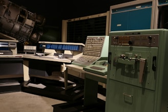 A vintage computer room featuring large, retro computers and control panels with numerous buttons and switches. The room includes a UNIVAC 1232 machine and several other large computing devices, arranged in a dimly lit space that emphasizes their mechanical and industrial design.