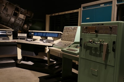 A vintage computer room featuring large, retro computers and control panels with numerous buttons and switches. The room includes a UNIVAC 1232 machine and several other large computing devices, arranged in a dimly lit space that emphasizes their mechanical and industrial design.