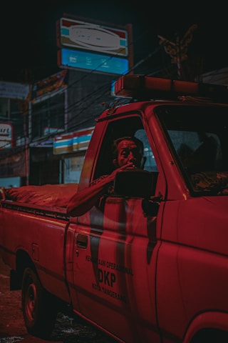 A red truck labeled with operational text is parked on a street at night. The scene is dimly lit, with neon lights from surrounding shops casting shadows and reflections. A person is visible inside the truck, looking out the window.