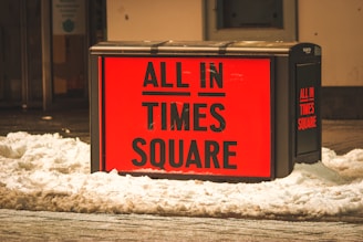 A bright red advertisement sign reads 'ALL IN TIMES SQUARE' and is surrounded by snow. The sign is mounted on a dark rectangular stand, situated in an urban area with a building in the background.