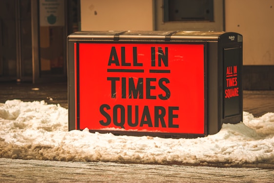 A bright red advertisement sign reads 'ALL IN TIMES SQUARE' and is surrounded by snow. The sign is mounted on a dark rectangular stand, situated in an urban area with a building in the background.