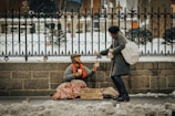 A person sitting on the ground, covered with a blanket, holding a cup and wearing warm clothes, receives a piece of bread from a passerby who is bending down. The background consists of a wrought-iron fence, snow-covered ground, and a stone wall. A sign with words is placed in front of the seated individual.