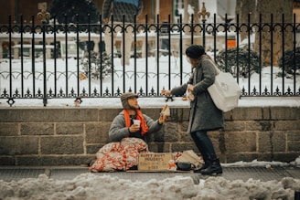 A person sitting on the ground, covered with a blanket, holding a cup and wearing warm clothes, receives a piece of bread from a passerby who is bending down. The background consists of a wrought-iron fence, snow-covered ground, and a stone wall. A sign with words is placed in front of the seated individual.