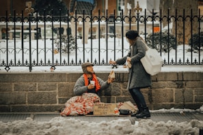 A person sitting on the ground, covered with a blanket, holding a cup and wearing warm clothes, receives a piece of bread from a passerby who is bending down. The background consists of a wrought-iron fence, snow-covered ground, and a stone wall. A sign with words is placed in front of the seated individual.