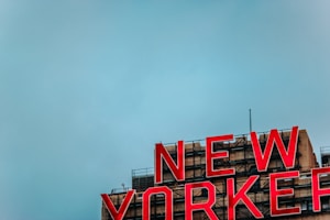 A large red neon sign spells out 'NEW YORKER' atop a building, with a clear blue sky in the background. The building has a beige facade with visible industrial elements and fixtures.