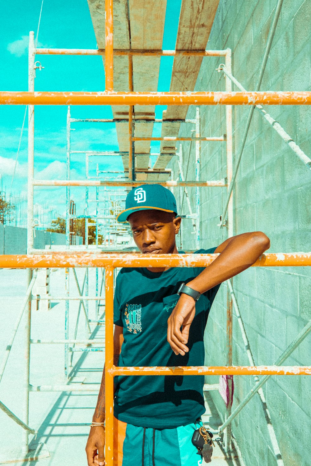 man in blue crew neck t-shirt and blue cap leaning on brown metal fence during