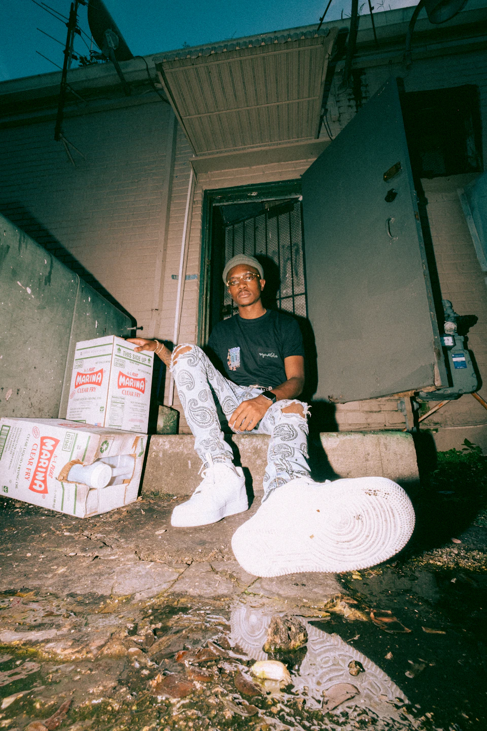 In the image, a young man is seated on a concrete ledge outside an open door, wearing a black t-shirt and white sneakers with a pair of ripped jeans. Behind him, there are two boxes stacked against the wall - one red and one green. The scene appears to be set in an urban environment, with power lines visible above the man's head.