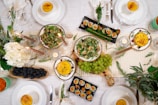 An elegant dining setting featuring a variety of dishes, including salads, sushi, and desserts. The table is beautifully decorated with candles, flowers, and greenery. There are plates with bright yellow desserts sprinkled with herbs, platters of sushi rolls, and bowls of fresh salad garnished with cranberries. Bright green grapes and blackberries are also part of the arrangement, adding to the vibrant and festive spread.