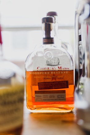 A close-up view of a bottle of whiskey with a dark brown cap, featuring a label with text and an illustration of a barrel. The bottle contains an amber-colored liquid and is positioned on a wooden surface with blurred objects in the background.