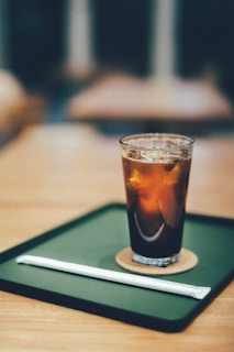 A glass filled with iced coffee or tea is placed on a green tray with a coaster beneath the glass. A wrapped straw lies next to it on the tray. The background is softly blurred, suggesting a warm, relaxed atmosphere.