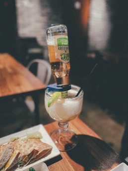 A large glass filled with a cocktail that has an overturned bottled drink inserted into it, finished with a wedge of lime. The setting appears to be a bar or restaurant, with warm lighting and wooden tables. There is also a plate of quesadillas with green garnish on the table.