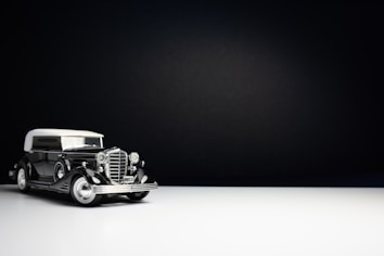 A vintage black and white car model from 1933 is positioned against a minimalist, dark background. The car has a glossy finish with distinctive features such as a large front grille, prominent headlights, and white-walled tires. The lighting highlights the car, casting a subtle reflection on the white surface below.