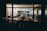 An office space viewed from the outside at night, featuring several desks, office chairs, and shelves filled with books and files. There are posters and photographs on the walls, and a person is seen working at a desk with a computer and lamp lit.