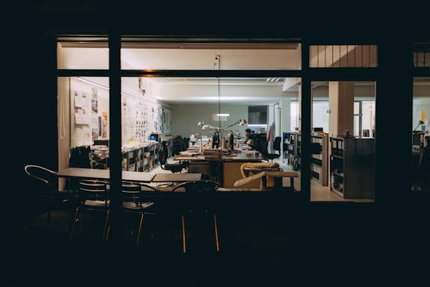 An office space viewed from the outside at night, featuring several desks, office chairs, and shelves filled with books and files. There are posters and photographs on the walls, and a person is seen working at a desk with a computer and lamp lit.