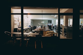 An office space viewed from the outside at night, featuring several desks, office chairs, and shelves filled with books and files. There are posters and photographs on the walls, and a person is seen working at a desk with a computer and lamp lit.