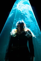 A person stands under a dramatic beam of blue light that appears to create an ethereal or mystical aura around them. The light accentuates swirling patterns, possibly smoke or mist, adding to the surreal atmosphere.