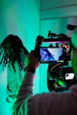 A person is being filmed with professional camera equipment in an environment illuminated by blue and green lighting. The focus is on the filming process, with the camera screen displaying the person being recorded.