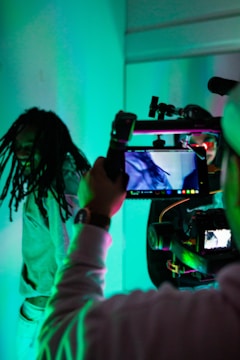 A person is being filmed with professional camera equipment in an environment illuminated by blue and green lighting. The focus is on the filming process, with the camera screen displaying the person being recorded.