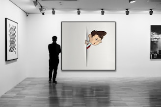 A person stands in a gallery space, observing a large, modern artwork that depicts an animated character peeking through a slit. The gallery is well-lit, with other artworks hanging on the white walls, and has a wooden floor.