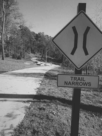 A black and white photograph of a pathway through a park area with trees lining both sides. A prominent sign in the foreground indicates that the trail narrows ahead. The path is winding and there are trees without leaves visible, suggesting a winter or early spring setting.