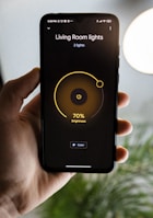 A hand is holding a smartphone displaying a smart lighting app. The screen shows controls for 'Living Room lights' with brightness set at 70%. The app has a minimalist design with a dark background and a central yellow circle indicating the brightness level. Part of a blurred plant and a bright round light are visible in the background.
