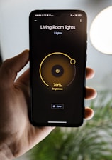 A hand is holding a smartphone displaying a smart lighting app. The screen shows controls for 'Living Room lights' with brightness set at 70%. The app has a minimalist design with a dark background and a central yellow circle indicating the brightness level. Part of a blurred plant and a bright round light are visible in the background.