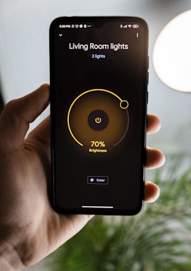 A hand is holding a smartphone displaying a smart lighting app. The screen shows controls for 'Living Room lights' with brightness set at 70%. The app has a minimalist design with a dark background and a central yellow circle indicating the brightness level. Part of a blurred plant and a bright round light are visible in the background.