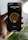A hand is holding a smartphone displaying a smart lighting app. The screen shows controls for 'Living Room lights' with brightness set at 70%. The app has a minimalist design with a dark background and a central yellow circle indicating the brightness level. Part of a blurred plant and a bright round light are visible in the background.