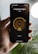 A hand is holding a smartphone displaying a smart lighting app. The screen shows controls for 'Living Room lights' with brightness set at 70%. The app has a minimalist design with a dark background and a central yellow circle indicating the brightness level. Part of a blurred plant and a bright round light are visible in the background.