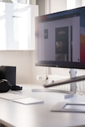A modern office workspace with a large computer monitor displaying a digital design. The monitor is positioned on a sleek white desk, accompanied by a wireless keyboard and mouse. There's a pair of headphones resting on the desk and some minimalistic shelving is visible in the background.