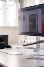 A modern office workspace with a large computer monitor displaying a digital design. The monitor is positioned on a sleek white desk, accompanied by a wireless keyboard and mouse. There's a pair of headphones resting on the desk and some minimalistic shelving is visible in the background.