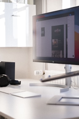 A modern office workspace with a large computer monitor displaying a digital design. The monitor is positioned on a sleek white desk, accompanied by a wireless keyboard and mouse. There's a pair of headphones resting on the desk and some minimalistic shelving is visible in the background.