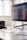 A modern office workspace with a large computer monitor displaying a digital design. The monitor is positioned on a sleek white desk, accompanied by a wireless keyboard and mouse. There's a pair of headphones resting on the desk and some minimalistic shelving is visible in the background.