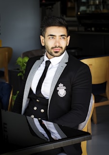 A stylish man in a classic black suit from Lumina Apparel, seated in a modern café with soft natural light.