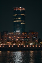 A high-rise building illuminated at night, with a cylindrical structure on top and warm lights glowing from the windows. The word 'Hyatt' is visible in white letters. The building is reflected in the water in the foreground.