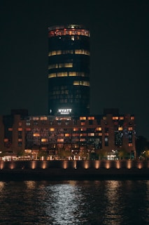 A high-rise building illuminated at night, with a cylindrical structure on top and warm lights glowing from the windows. The word 'Hyatt' is visible in white letters. The building is reflected in the water in the foreground.