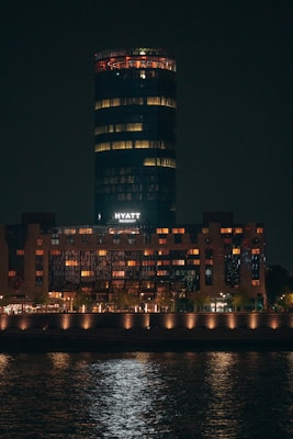 A high-rise building illuminated at night, with a cylindrical structure on top and warm lights glowing from the windows. The word 'Hyatt' is visible in white letters. The building is reflected in the water in the foreground.