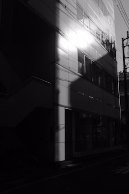 A building exterior with a modern facade reflects bright sunlight on its surface. The light creates dramatic contrasts with shadows, highlighting the structure's geometric design. Several windows are visible, and the lines of the building are crisp and angular. A street leading to the building is partially visible with bicycles parked nearby.