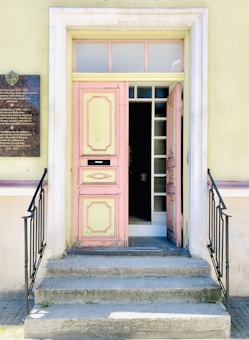 A pastel-colored doorway with an ornate frame leads into a building. The door is partially open, revealing a dark interior. Steps with metal railings lead up to the entrance. A plaque is mounted on the wall to the left of the door.