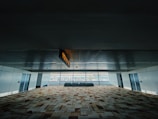 A spacious indoor corridor with patterned carpet flooring, surrounded by white walls and ceiling. Large windows line the far end, allowing natural light to brighten the space. A ceiling sign is directing towards exits, and a small row of plants is visible in front of the windows.