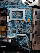 A detailed close-up of a computer motherboard with a variety of electronic components, including microchips, resistors, and connectors. The board is mostly blue with some metallic and black areas, displaying numerous intricate patterns and circuits.