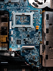 A detailed close-up of a computer motherboard with a variety of electronic components, including microchips, resistors, and connectors. The board is mostly blue with some metallic and black areas, displaying numerous intricate patterns and circuits.