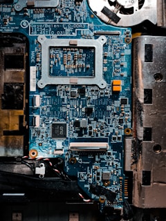 A detailed close-up of a computer motherboard with a variety of electronic components, including microchips, resistors, and connectors. The board is mostly blue with some metallic and black areas, displaying numerous intricate patterns and circuits.