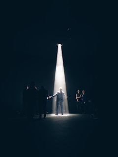 A person stands dramatically in a bright spotlight, surrounded by darkness with a group of other people partially visible in the shadows. The scene suggests a theatrical or performance setting with a focus on the central figure.