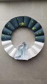 A decorative wreath made of individual segments that transition in color from dark blue to white. The wreath is adorned with small green star stickers and has a larger yellow star at the top. At the center of the wreath is a paper cutout depicting a nativity scene with two figures and animals, symbolizing a traditional Christmas theme.