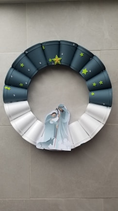 A decorative wreath made of individual segments that transition in color from dark blue to white. The wreath is adorned with small green star stickers and has a larger yellow star at the top. At the center of the wreath is a paper cutout depicting a nativity scene with two figures and animals, symbolizing a traditional Christmas theme.