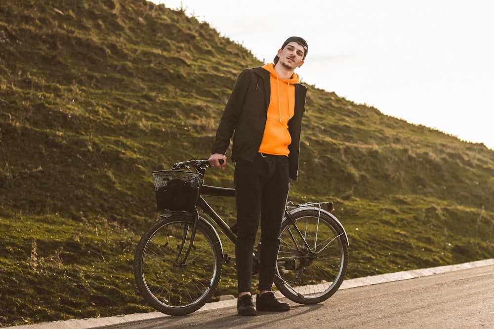 man in black jacket and orange shirt standing beside black bicycle