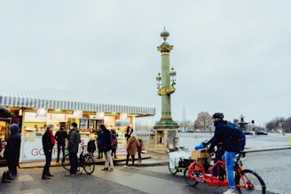 A group of people is gathered in front of a street food stall, possibly selling sandwiches and other snacks. The stall has a striped awning, and people appear to be queuing. A cyclist on a red bicycle is passing by, carrying goods in a cargo box on the back. A decorative street lamp stands nearby in an urban setting.